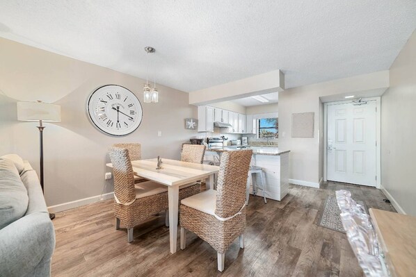 A modern living room with a dining table and chairs, a kitchen in the background, and a large wall clock. - Seaside Beach and Racquet 5216 | BeachFront + Pool (Orange Beach)