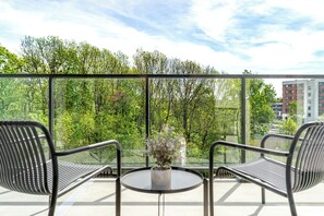 A balcony with two chairs and a table, overlooking a lush green area and a building.