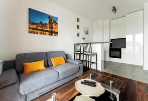 A modern living room with a grey sofa, a painting of a cityscape, a dining area with chairs, and a kitchen with white cabinets.
