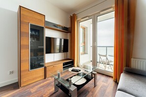 A modern living room with a glass coffee table, wooden cabinet, flat-screen TV, and a balcony with a view.