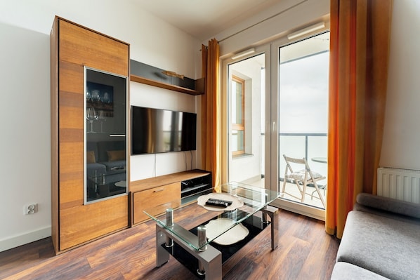 A modern living room with a glass coffee table, wooden cabinet, flat-screen TV, and a balcony with a view.