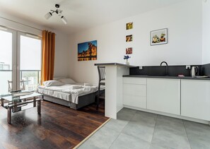 A modern room with a bed, a glass table, a chair, a kitchenette, and a city view.