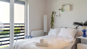 Een slaapkamer met een bed, een radiator, een klein tafeltje, een blauwe stoel, een balkon met uitzicht, en een wandplank met decoratieve objecten.