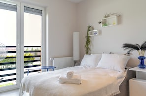 Ein Schlafzimmer mit Bett, Heizkörper, einem kleinen Tisch, einem blauen Stuhl, einem Balkon mit Blick und einem Wandregal mit Dekoration.