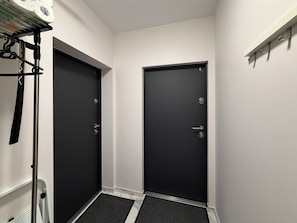 Ein schmaler Flur mit zwei schwarzen Türen, einer weißen Wand und einem Regal mit Gegenständen auf der rechten Seite.