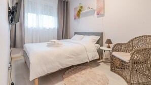 Ein ordentlich eingerichtetes Schlafzimmer mit einem Bett, einem Nachttisch, einem Korbstuhl und einem an der Wand montierten Fernseher.
