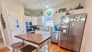 A kitchen with white cabinets, a wooden island with stools, a stainless steel refrigerator, and a window with a view. - Coastal 3BR Cottage w/ Pool Access & EV Charger | The Rookery 900 Fort Morgan (Gulf Shores)