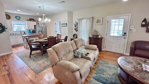 A living room with a beige sofa, a dining area with a wooden table and chairs, a kitchen with white cabinets, and a ceiling fan. - Coastal 3BR Cottage w/ Pool Access & EV Charger | The Rookery 900 Fort Morgan (Gulf Shores)
