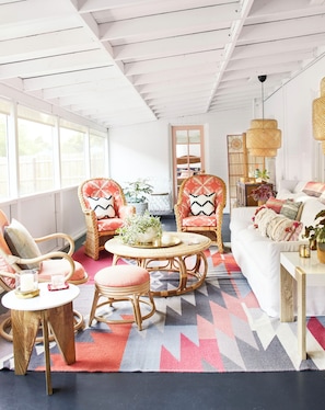 Une pièce spacieuse avec une grande fenêtre, des meubles en osier, un tapis à motifs et un canapé blanc.