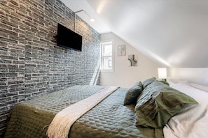 Een slaapkamer met een bakstenen muur, een bed met groen en wit beddengoed, een flatscreen-televisie aan de muur en een raam dat natuurlijk licht binnenlaat.