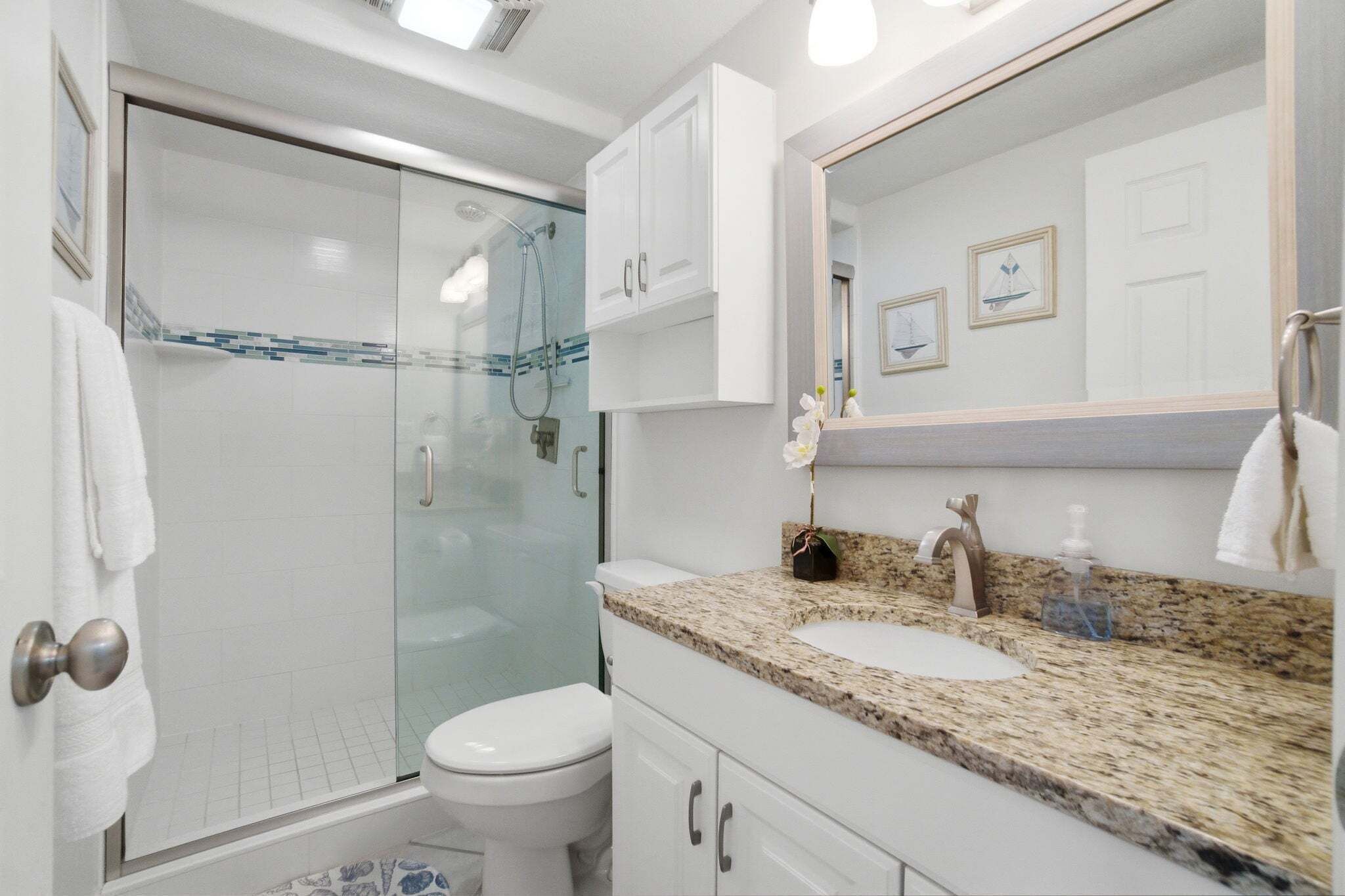 Une salle de bain avec une douche à parois de verre, des toilettes blanches, un lavabo sur un plan de travail en granit et un grand miroir.