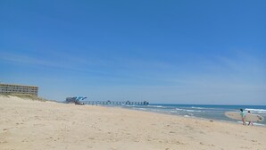 Een strand met een pier die zich uitstrekt over de oceaan, een gebouw met een trap aan de linkerkant en een persoon met een surfplank aan de rechterkant.