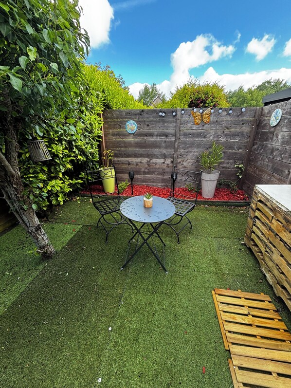 Un patio trasero con una mesa redonda y sillas, rodeado de césped artificial, plantas en macetas y una valla de madera con detalles decorativos.