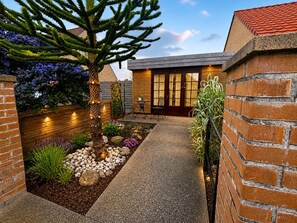 Una casa moderna con fachada de ladrillo, una terraza de madera y un jardín con diversas plantas y un árbol.