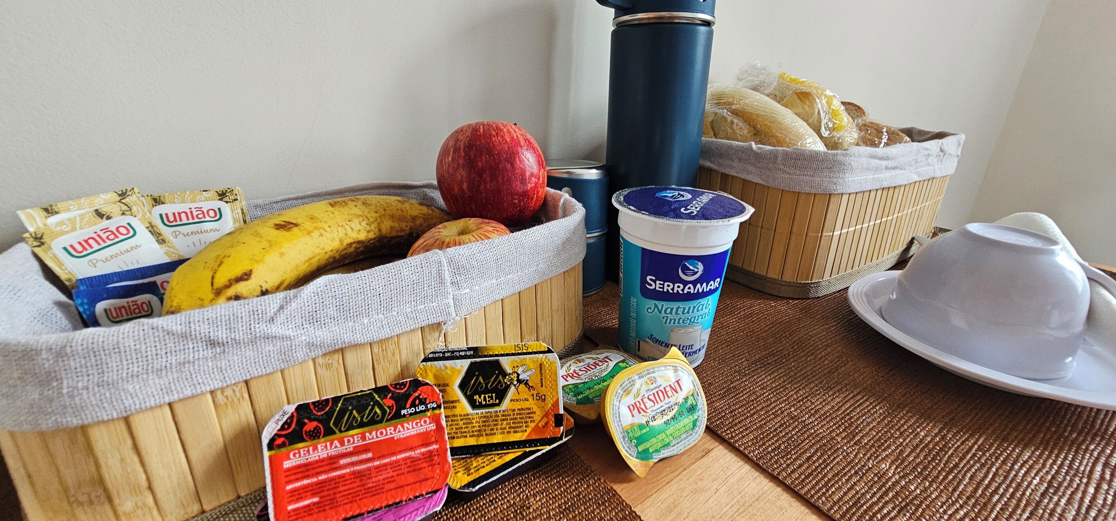 A breakfast setting with a basket containing a banana, an apple, and bread, a thermos, a cup of yogurt, and a plate with a teapot.