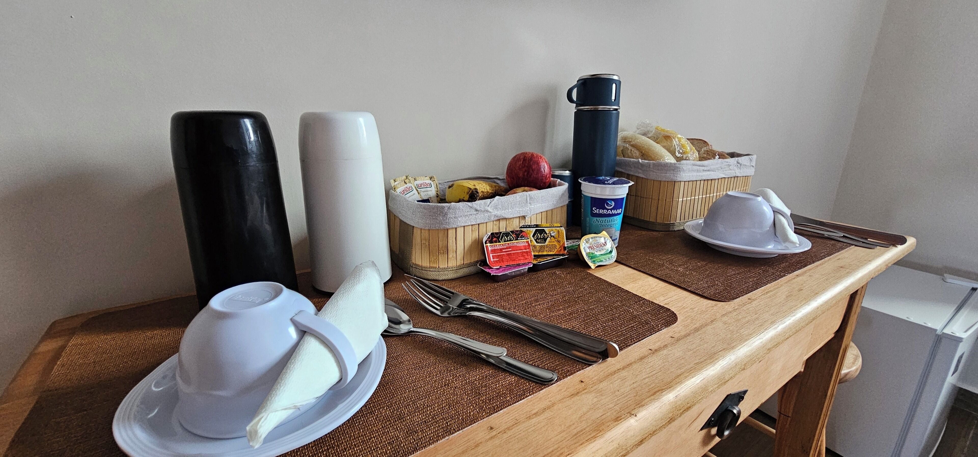 A breakfast setup with a basket of fruits, a thermos, a yogurt cup, and a plate with a napkin.