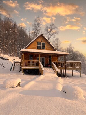 Una cabaña de madera con porche y escalones que conducen a la entrada, rodeada de nieve y árboles.