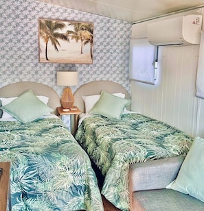 Ein Zimmer mit zwei Betten, einer Wand-Klimaanlage, einem gerahmten Bild von Palmen und einem kleinen Tisch mit einer Lampe.