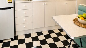 Une petite cuisine équipée comprenant un réfrigérateur, une fenêtre avec vue, une petite table avec un saladier de fruits et un sol à carreaux.