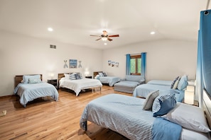 A bedroom with multiple beds, a ceiling fan, wooden flooring, and a window with blue curtains.