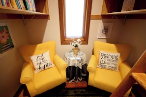 Un rincón de lectura acogedor con dos sillones amarillos, una mesita con un florero y un ventanal con luz natural.