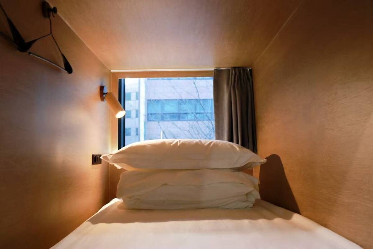 Et lille, smalt hotelværelse med en enkelt seng, et vindue med gardiner og en lampe på væggen.