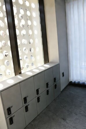 A modern room with a light-colored cabinet with numbered compartments, a wall-mounted light fixture, and a sheer curtain. - L YOUNG  Inn Capsule Hotel  Myeongdong (Seoul)
