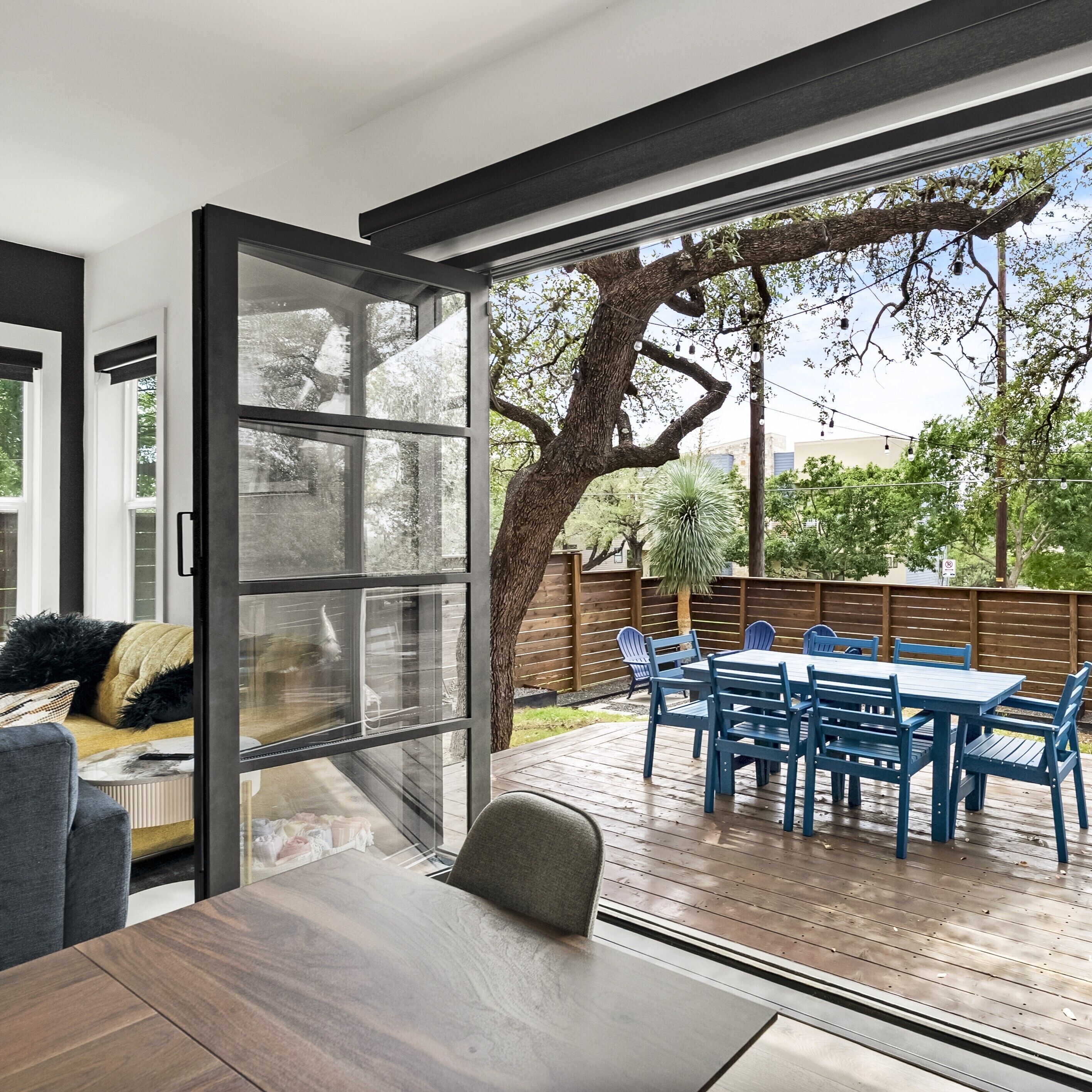 A modern patio with a wooden deck, a dining table with blue chairs, and a large glass door leading to an outdoor area with trees and a fence.