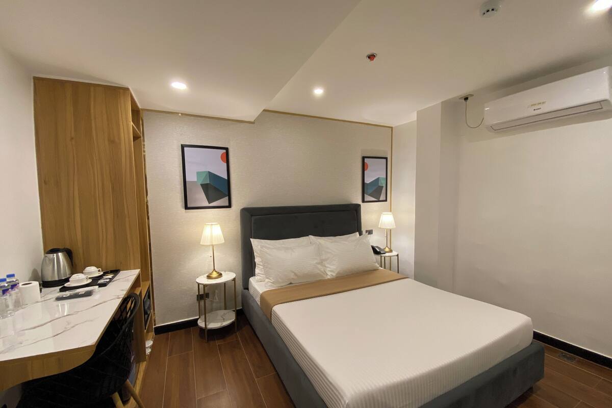 Una habitación de hotel con cama, mesitas de noche, escritorio con silla, cafetera y dos aires acondicionados instalados en la pared.