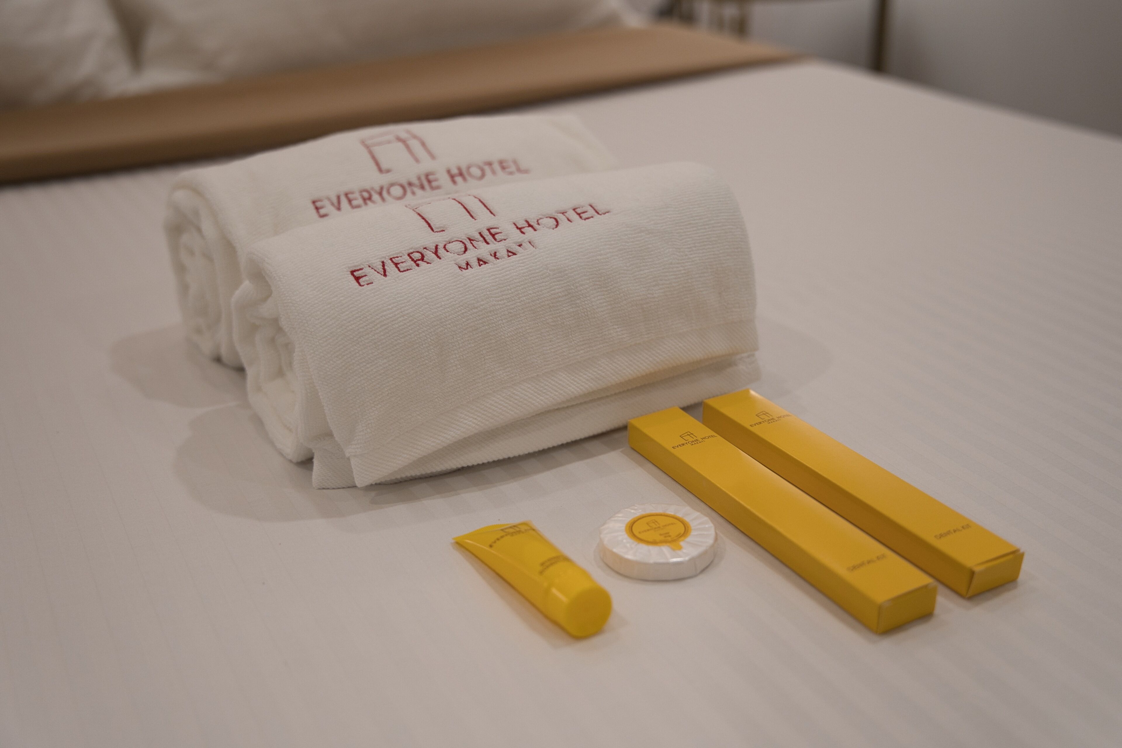 A neatly folded white towel with 'EVERYONE HOTEL' text, two yellow boxes, and a small white item on a bed.