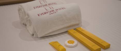 Une serviette blanche, soigneusement pliée, avec l’inscription « EVERYONE HOTEL », deux boîtes jaunes et un petit objet blanc posé sur un lit.