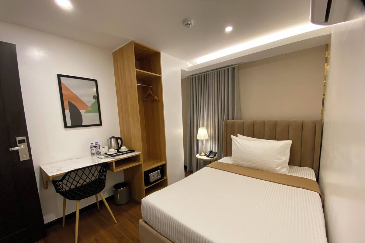 Habitación de hotel con cama, escritorio con silla, perchero, lámpara y un cuadro en la pared.