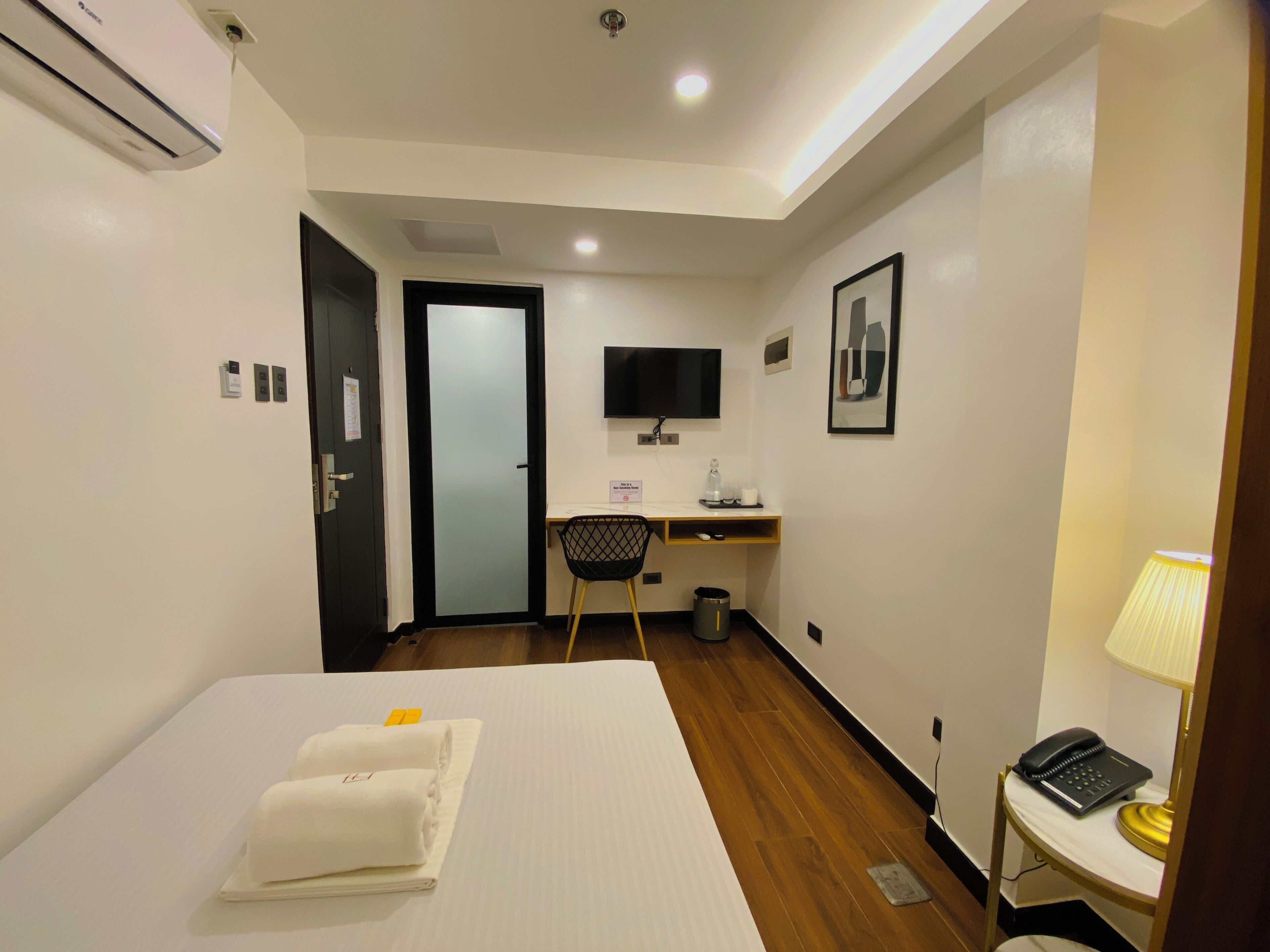 A hotel room with a bed, a desk, a chair, a TV, a lamp, and a trash bin.