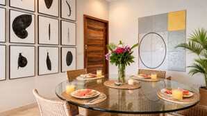 Een eethoek met een glazen tafel, omringd door rieten stoelen, een houten deur en kunstwerken in lijsten aan de muur.