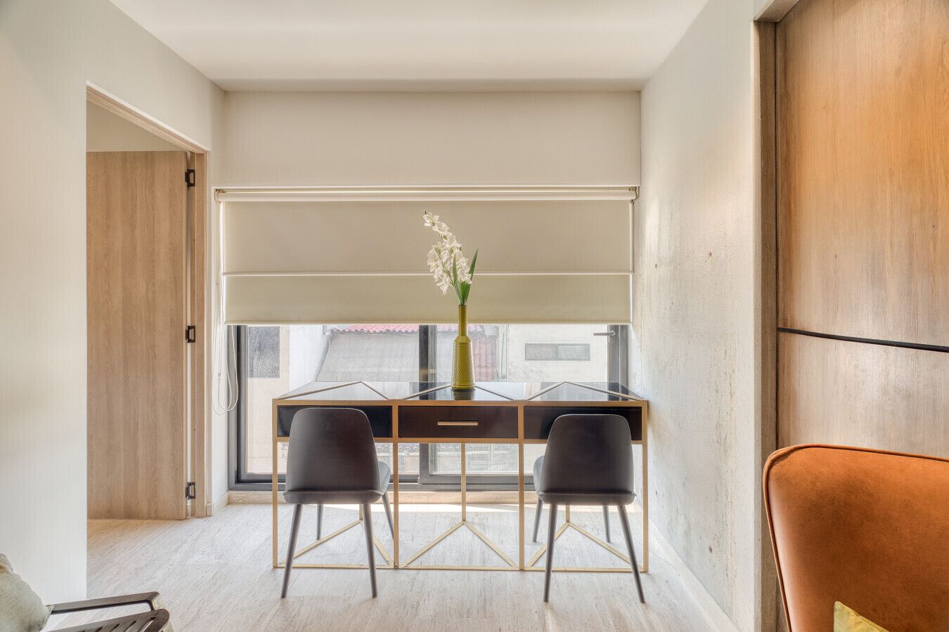 A modern office space with a desk, two chairs, a vase with flowers, and a view through a large window.