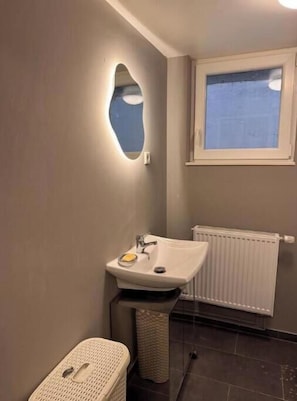 Ein Badezimmer mit einem Standwaschbecken, einem kleinen Fenster, einer Heizung und einem Korbwäscheschrank.