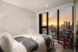 A bedroom with a large bed, a city view through floor-to-ceiling windows, and a red chair. - Modern City Gem 3BR | Pool, Gym and 2 Free Parking (Southbank)