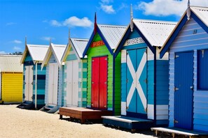 A row of colorful beach huts with numbered doors, each with a small wooden bench in front. - Modern City Gem 3BR | Pool, Gym and 2 Free Parking (Southbank)