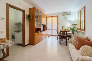 Ein Wohnzimmer mit einer Couch, einem Esstisch mit Stühlen, einer Küchenzeile mit Schränken und einer Glasschiebetür, die auf einen Balkon führt.