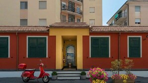 Una scooter roja estacionada frente a un edificio con contraventanas verdes, una entrada amarilla y macetas con flores.