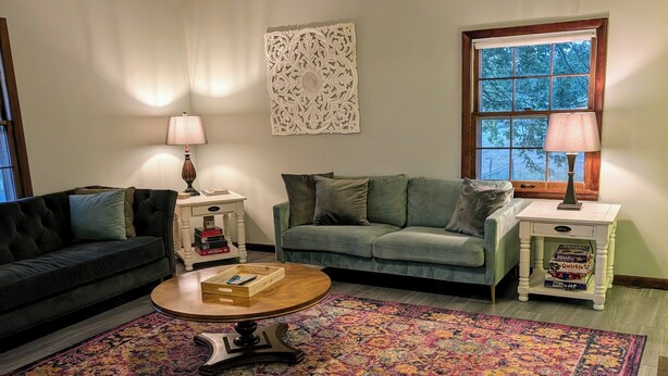 Uma sala de estar com ventilador de teto, duas luminárias, tapete estampado, sofá verde e mesa de centro.