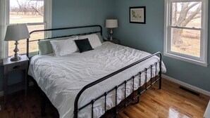 Quarto com cama, mesa de cabeceira, abajur, ventilador de teto e duas janelas.