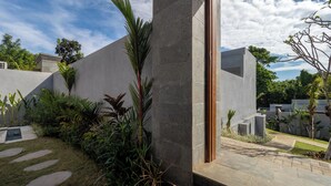 A modern building with a stone facade, a wooden door, and a garden with various plants. - Prawara Uluwatu (Pecatu)