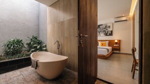 A modern bathroom with a freestanding bathtub, a wooden door, and a view into a bedroom with a bed and a nightstand. - Prawara Uluwatu (Pecatu)
