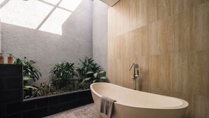 A modern bathroom with a freestanding bathtub, a showerhead, and a built-in planter with greenery. - Prawara Uluwatu (Pecatu)