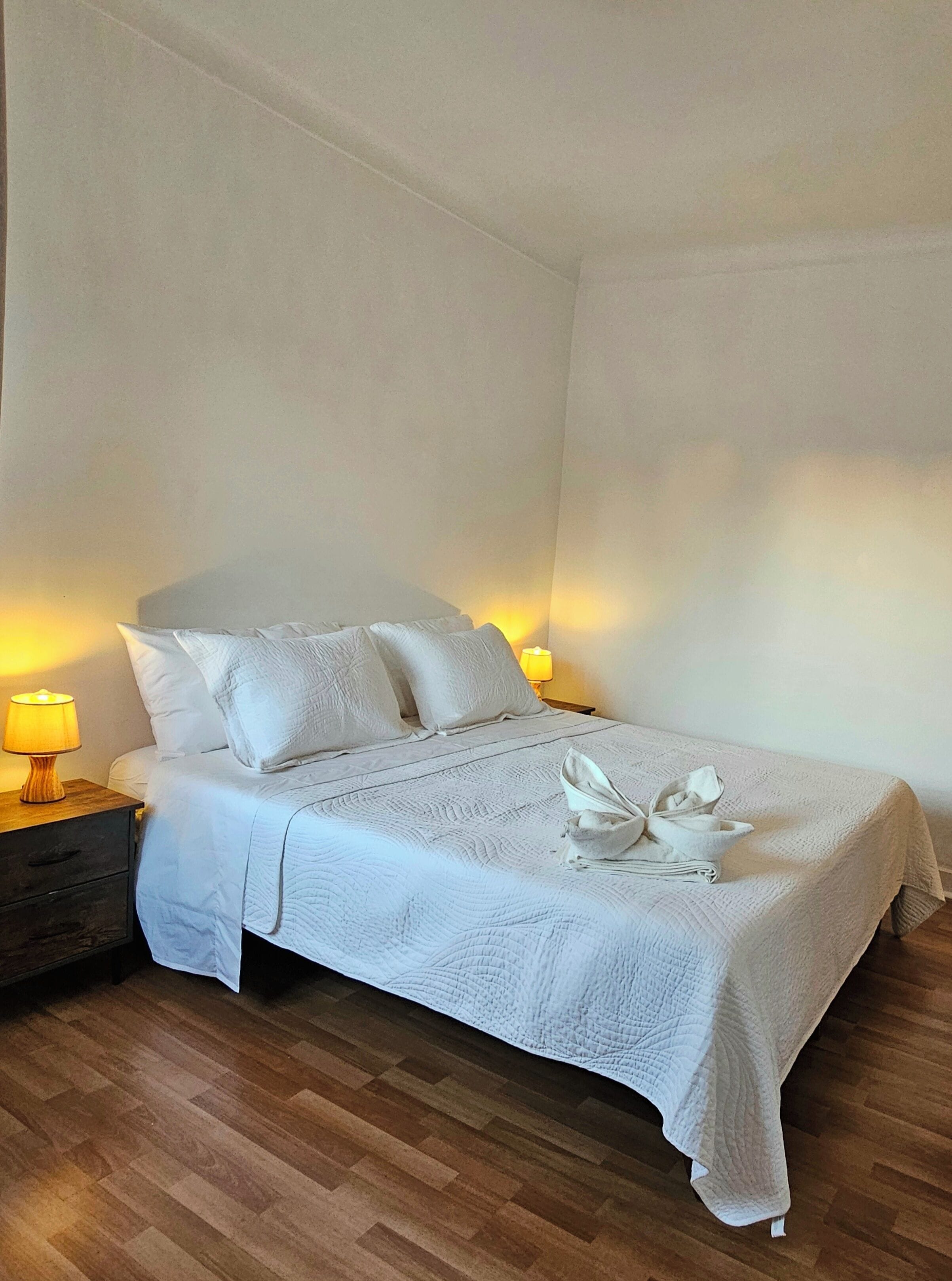 A neatly made bed with a white duvet and pillows, a wooden nightstand with a lamp, and a wooden floor.