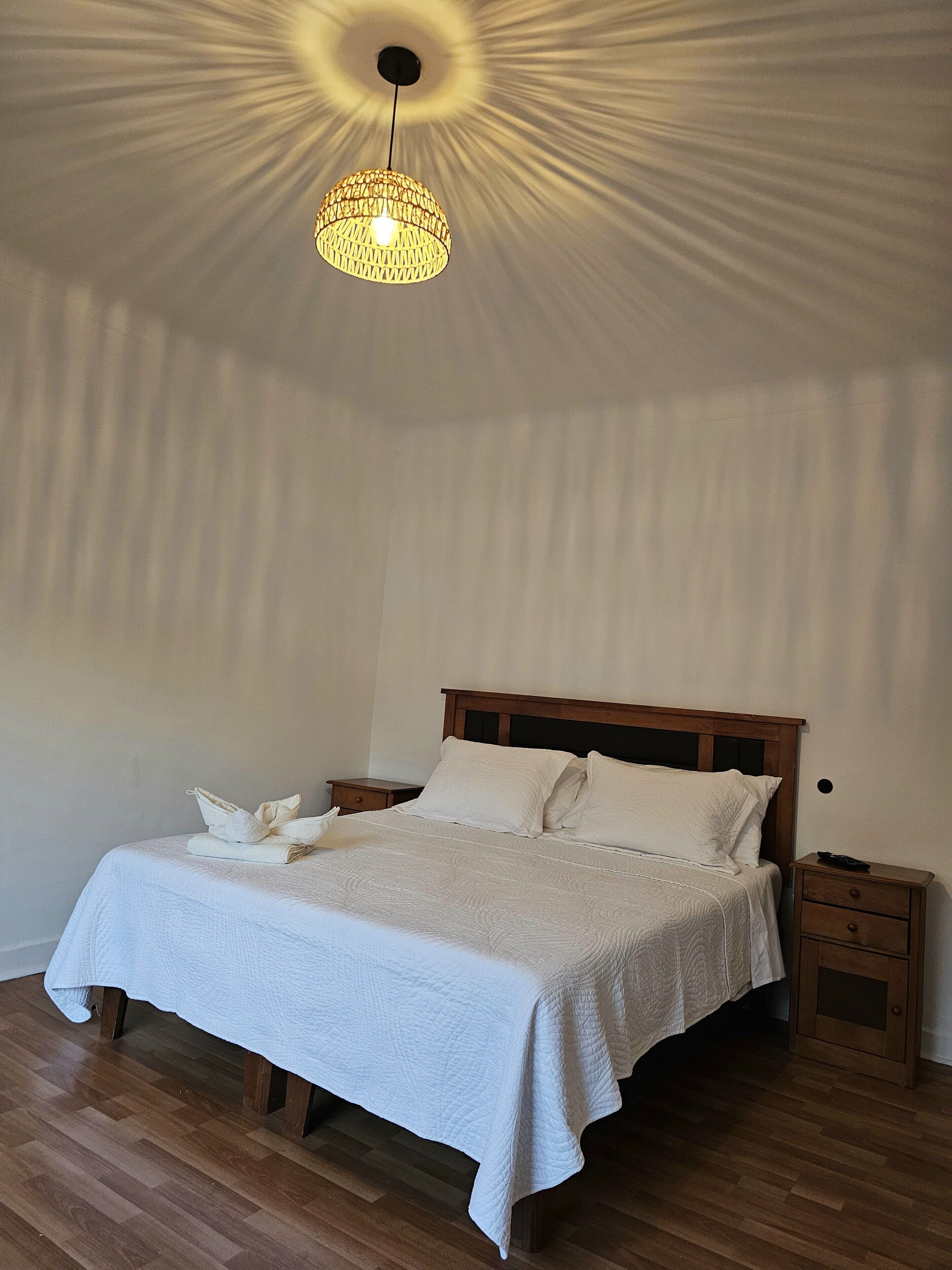 A neatly made bed with a white bedspread and pillows, a wooden nightstand with a drawer, and a hanging light fixture.