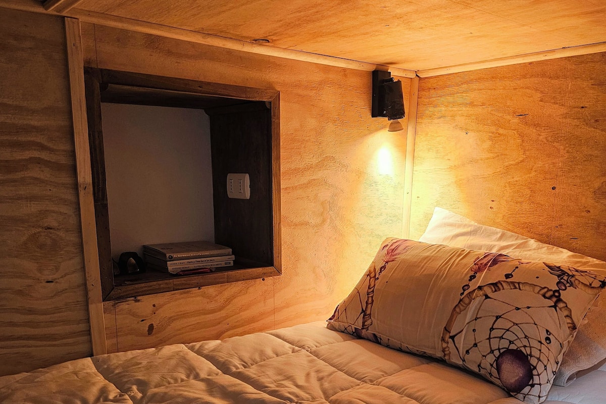 A single bed with a quilt, a bedside lamp, and a small shelf with books.