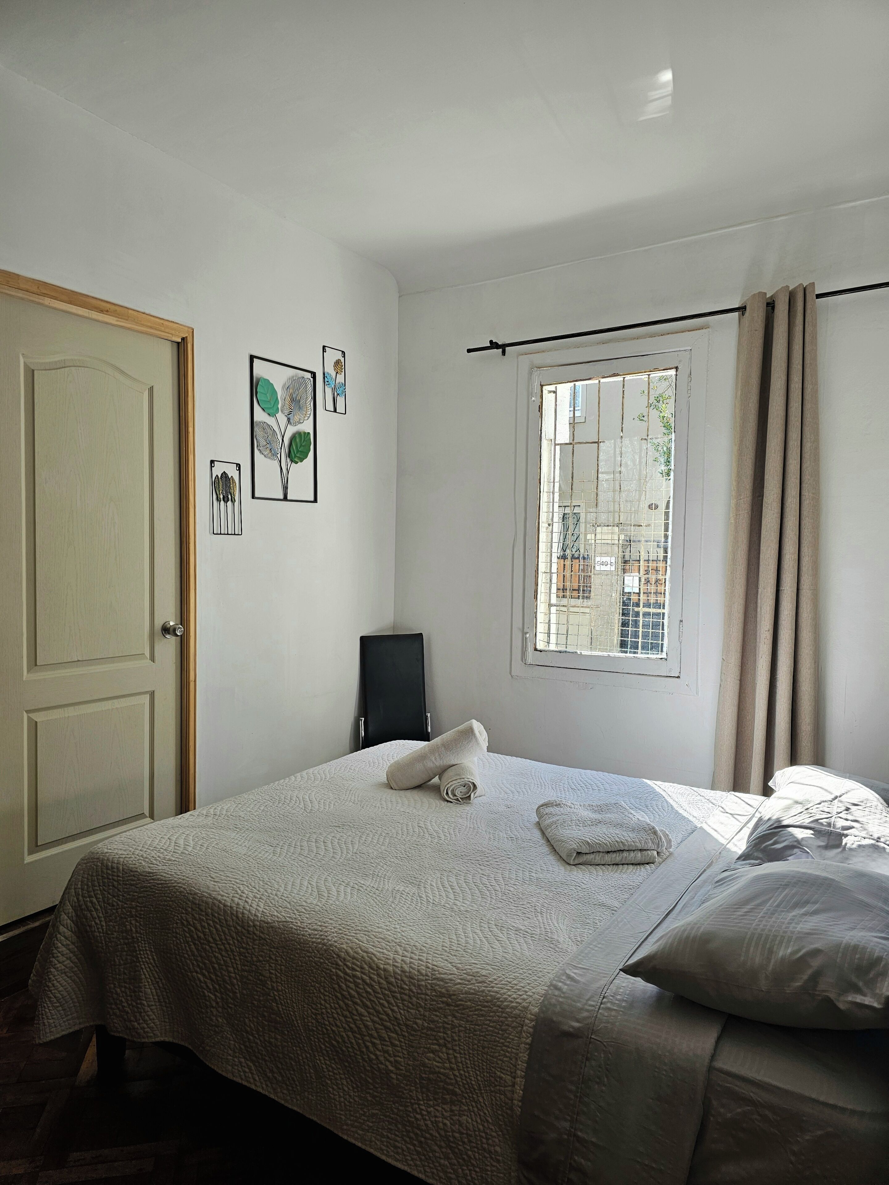 A neatly made bed with a rolled towel, a black chair, a window with curtains, and a door.
