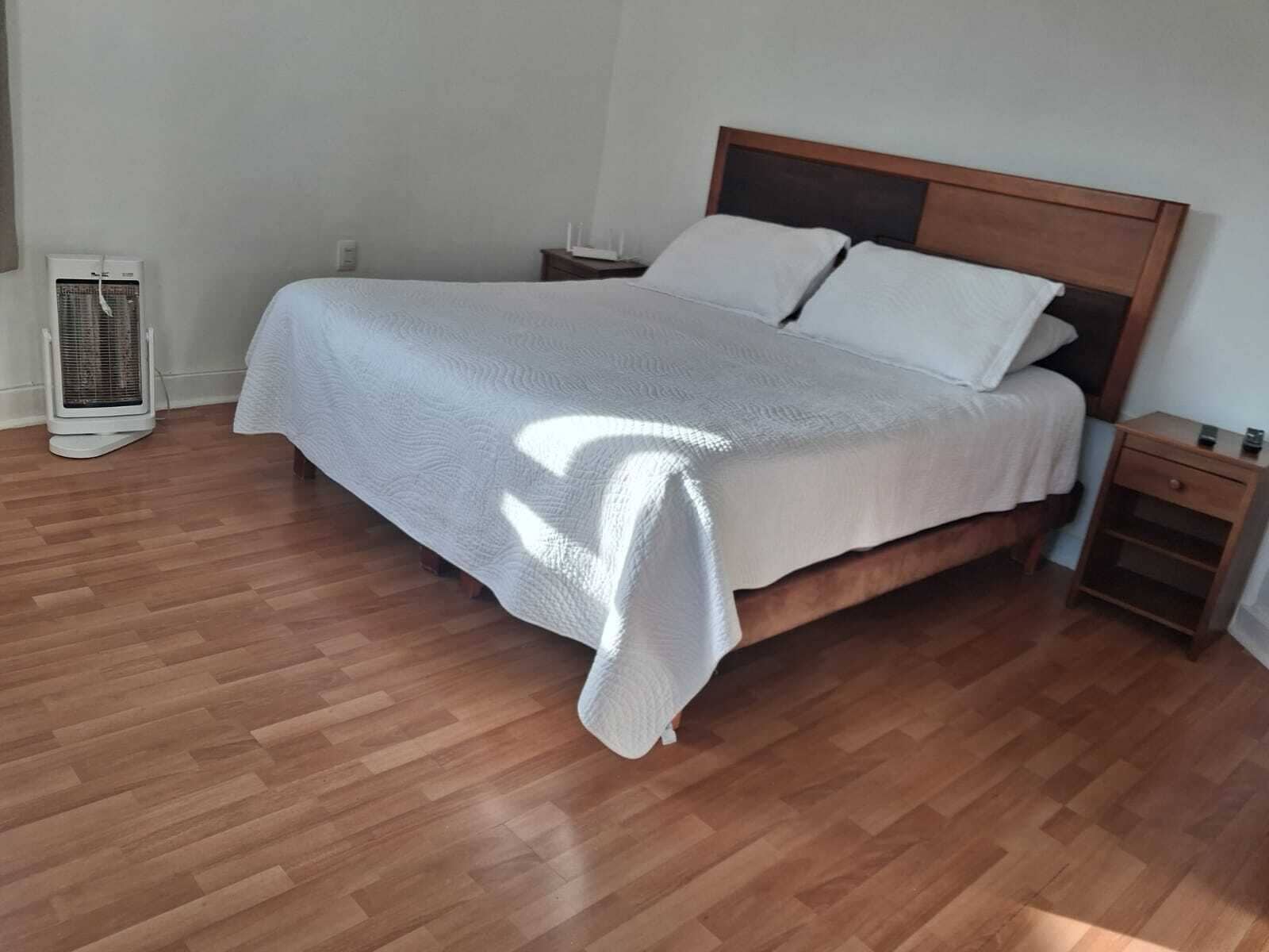 A neatly made bed with a wooden headboard, a bedside table with a lamp, and a standing heater.